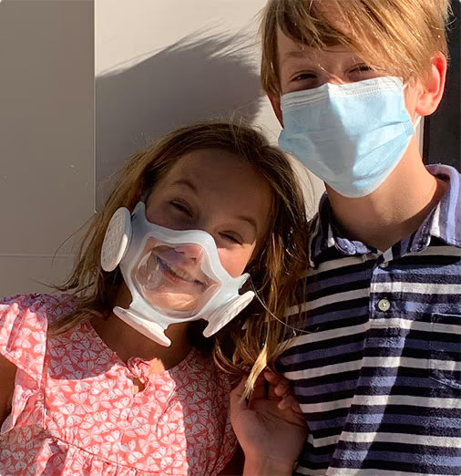 Young girl wearing her OmniMask and smiling at the camera while standing next to her brother in surgical mask.