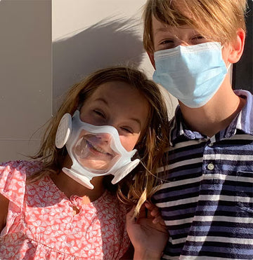 Young girl wearing her OmniMask and smiling at the camera while standing next to her brother in surgical mask.